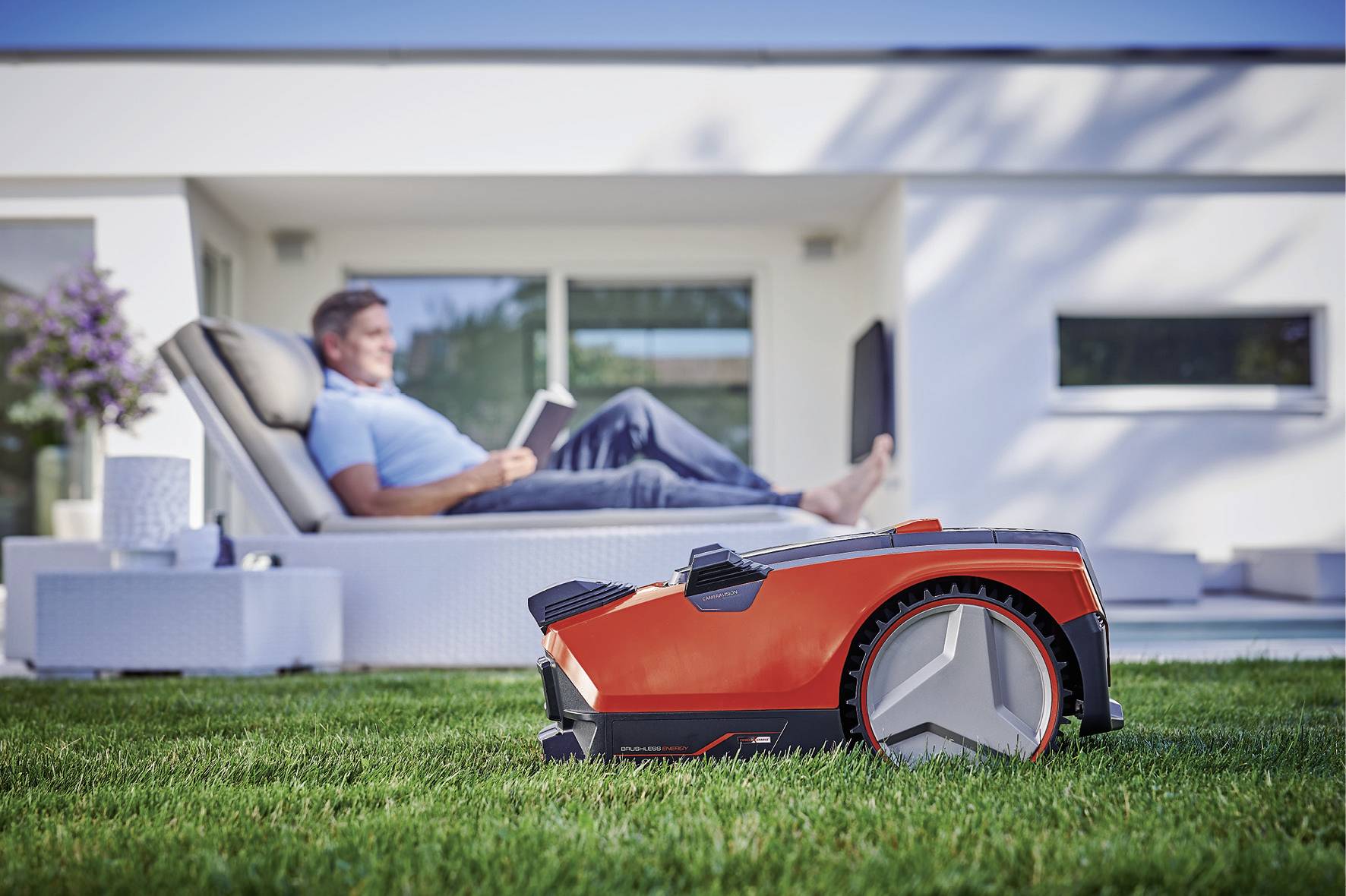 A red robotic lawnmower is cutting the grass in the foreground, while a person sits reading a gardening book on a lounger in the background.