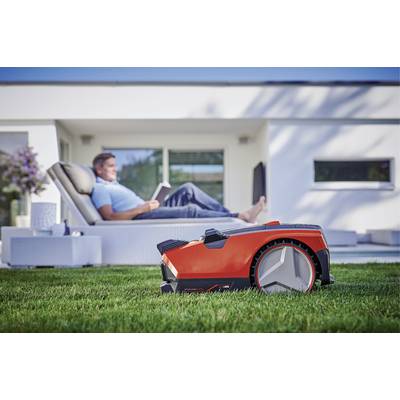A red robotic lawnmower is cutting the grass in the foreground, while a person sits reading a gardening book on a lounger in the background.