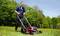 A man is mowing the lawn with an electric lawnmower on a meadow on a sunny day, surrounded by trees.