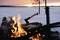 A campfire burns in front of a snow-covered landscape during sunset. An axe is stuck in the wood, fish grilling on the barbecue.