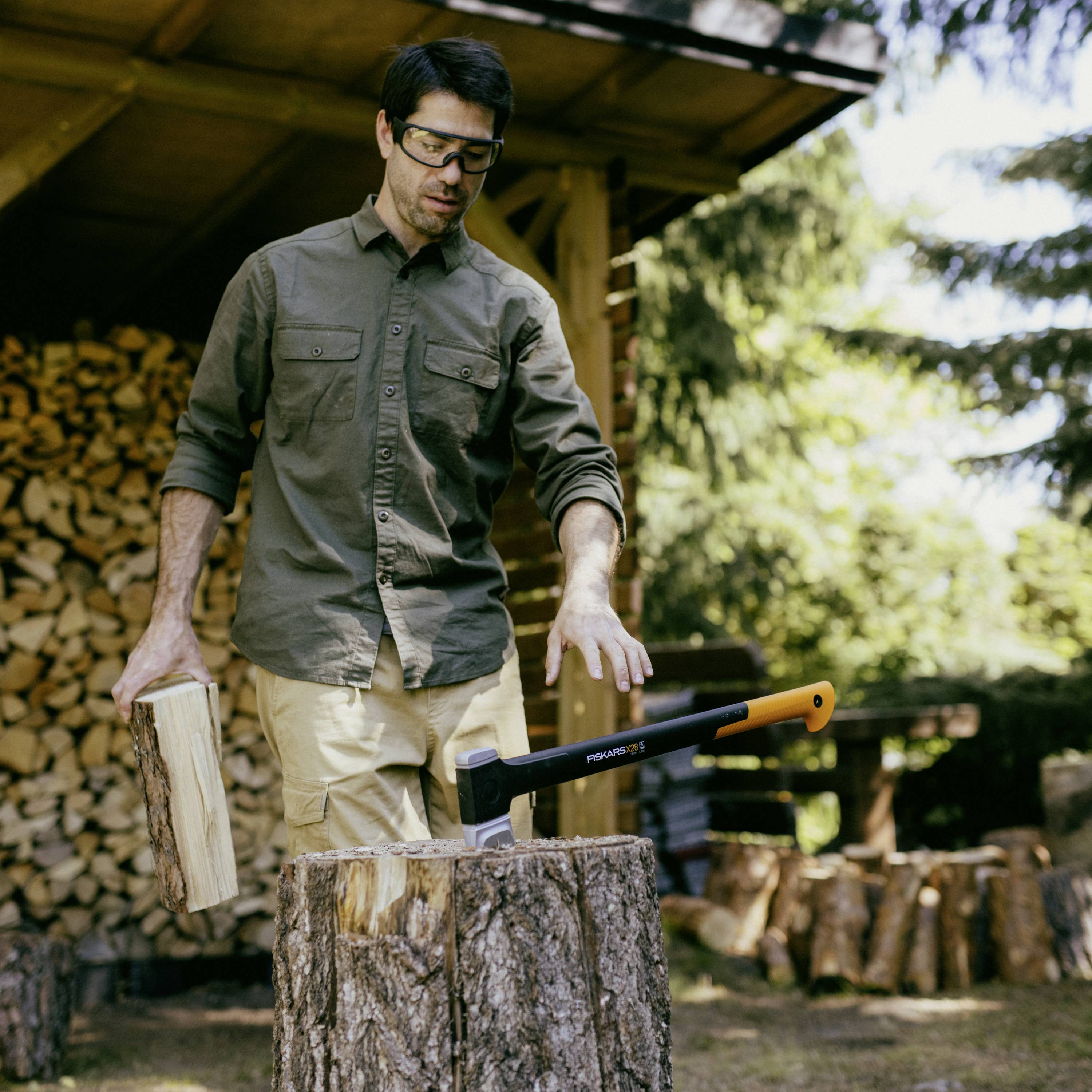 A person is standing outdoors in front of a woodpile, splitting a log with an axe. They are wearing safety glasses.