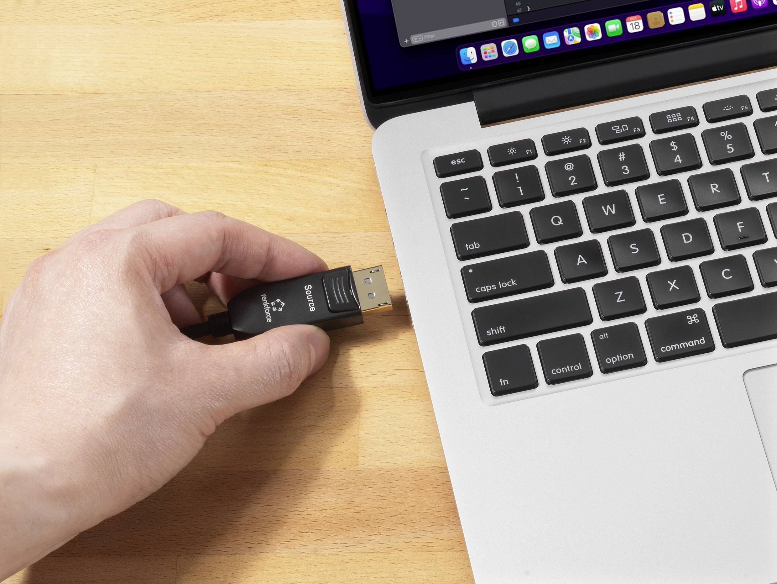 A hand is inserting a USB stick into a laptop on a wooden table. The laptop screen shows partially open programmes.