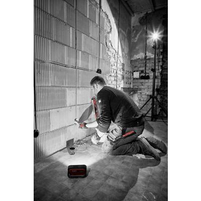 A worker kneels in front of a wall in an unfinished building and works on the wiring with tools. A construction floodlight illuminates the room.