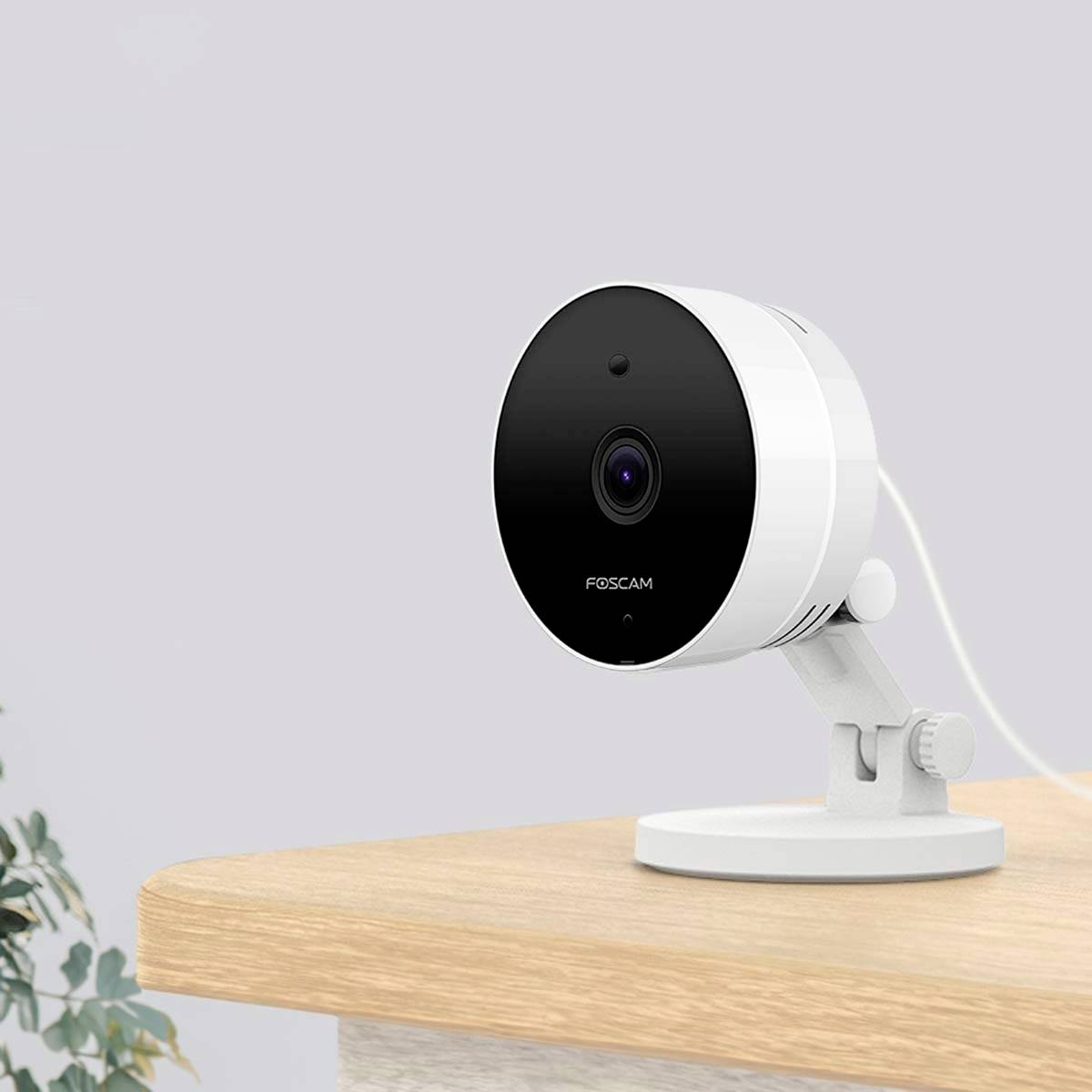 Security camera on a wooden table, showing the front view with lens. Model name visible on the front of the camera.