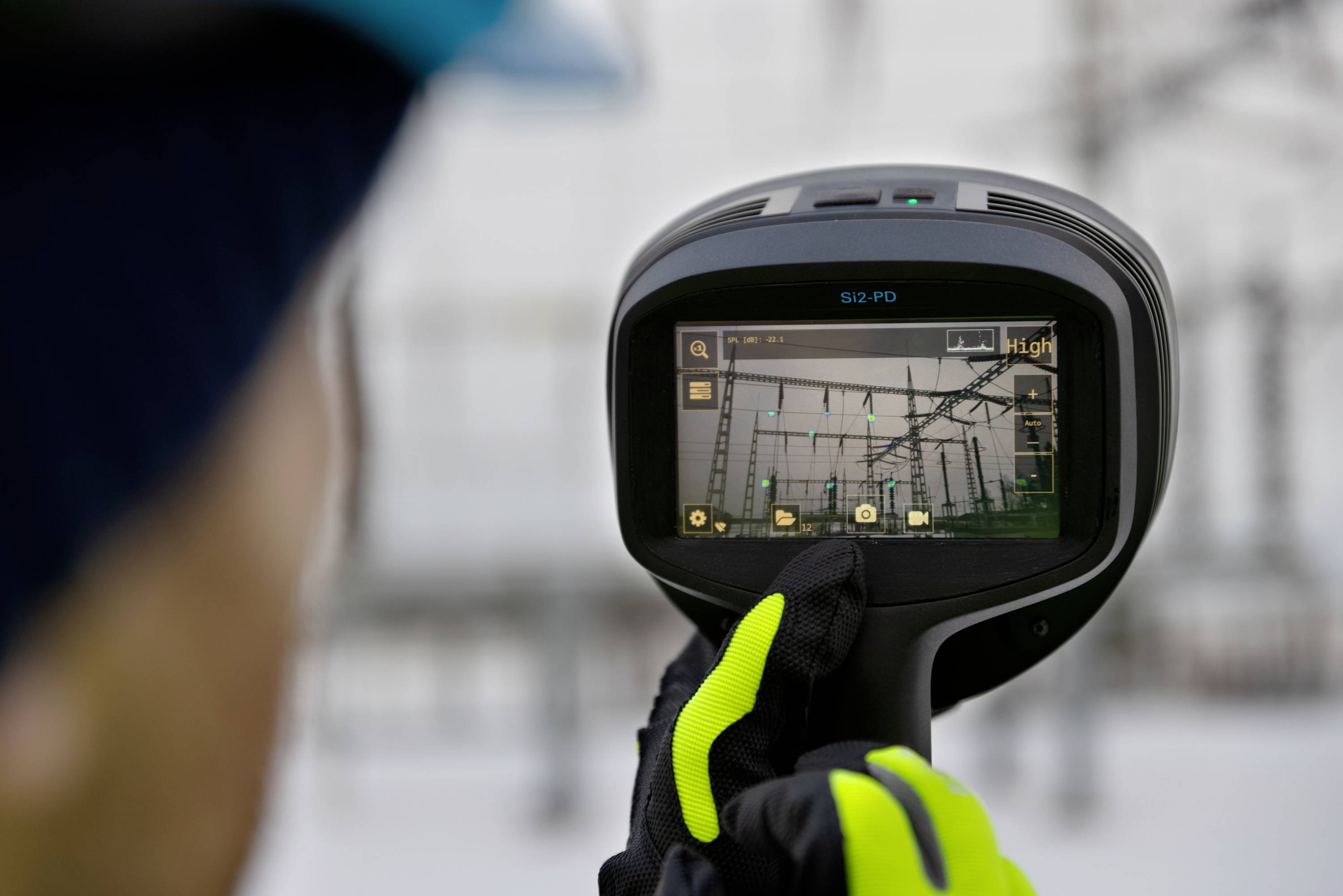 A person is holding an infrared camera device while inspecting part of an industrial facility. The display shows a thermal imaging view.