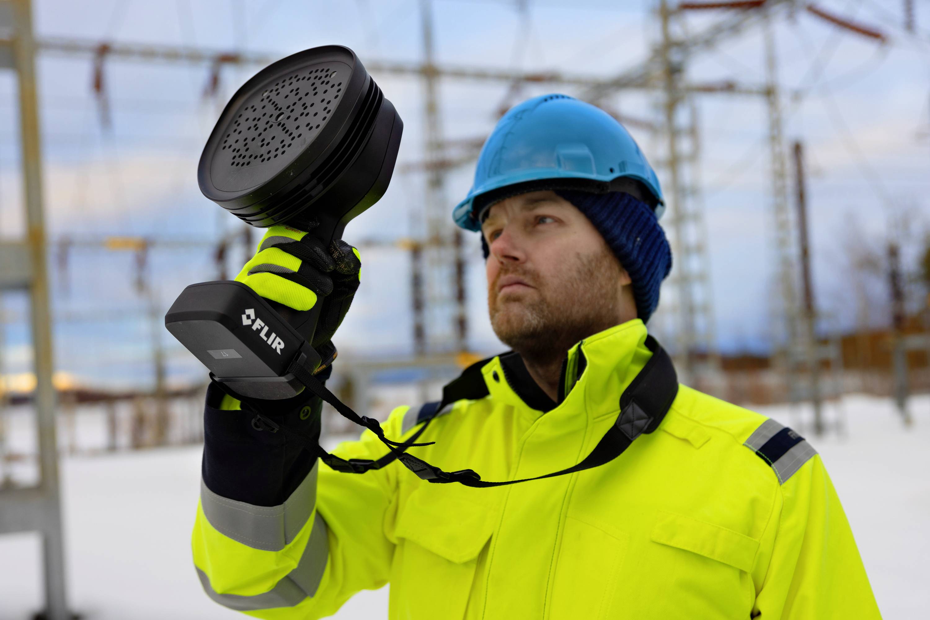 A man in protective workwear is holding a technical device, surrounded by electrical installations outdoors.