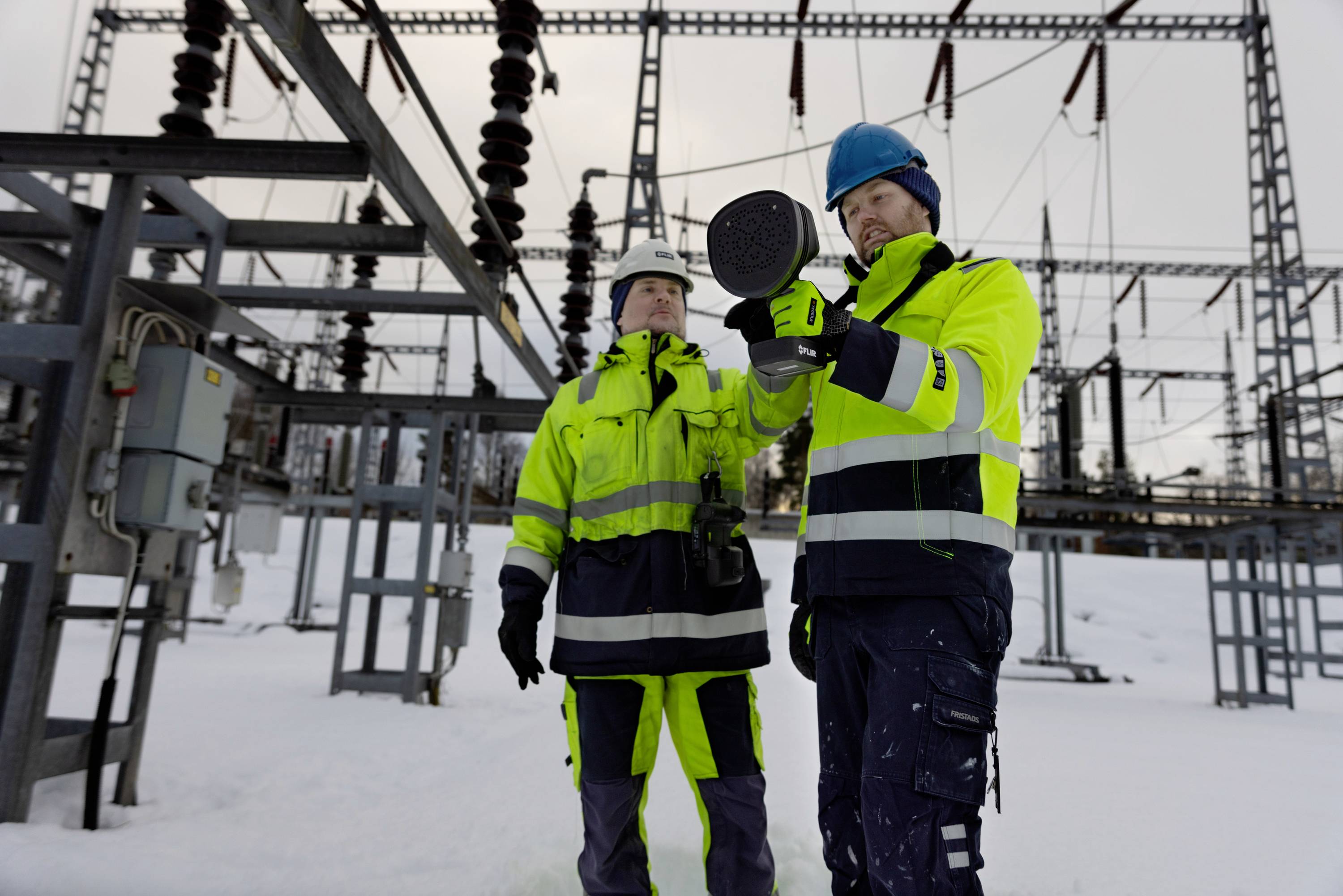 Two workers in safety clothing inspect technical equipment in a substation during winter, surrounded by snow and steel structures.