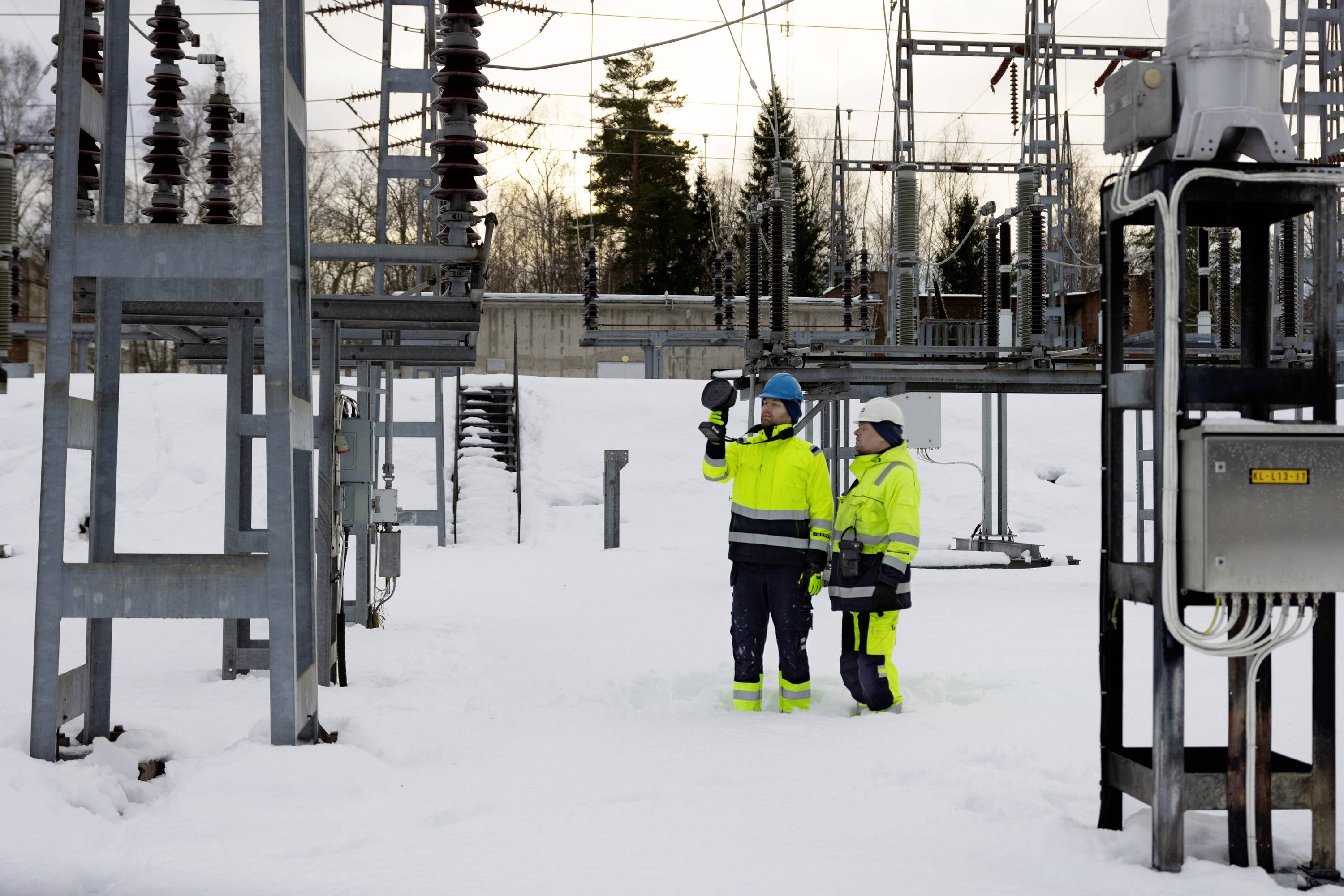 'Two people in work attire are checking electrical systems in a snow-covered outdoor area of a substation, surrounded by metal structures.'
