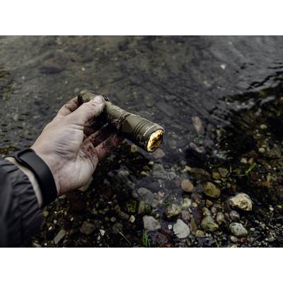 A hand holds a dirty torch over a flat, stony riverbed. The torch is switched on.