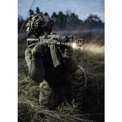A person in camouflage clothing kneels, holding a rifle with an attached torch outdoors at dusk or night.