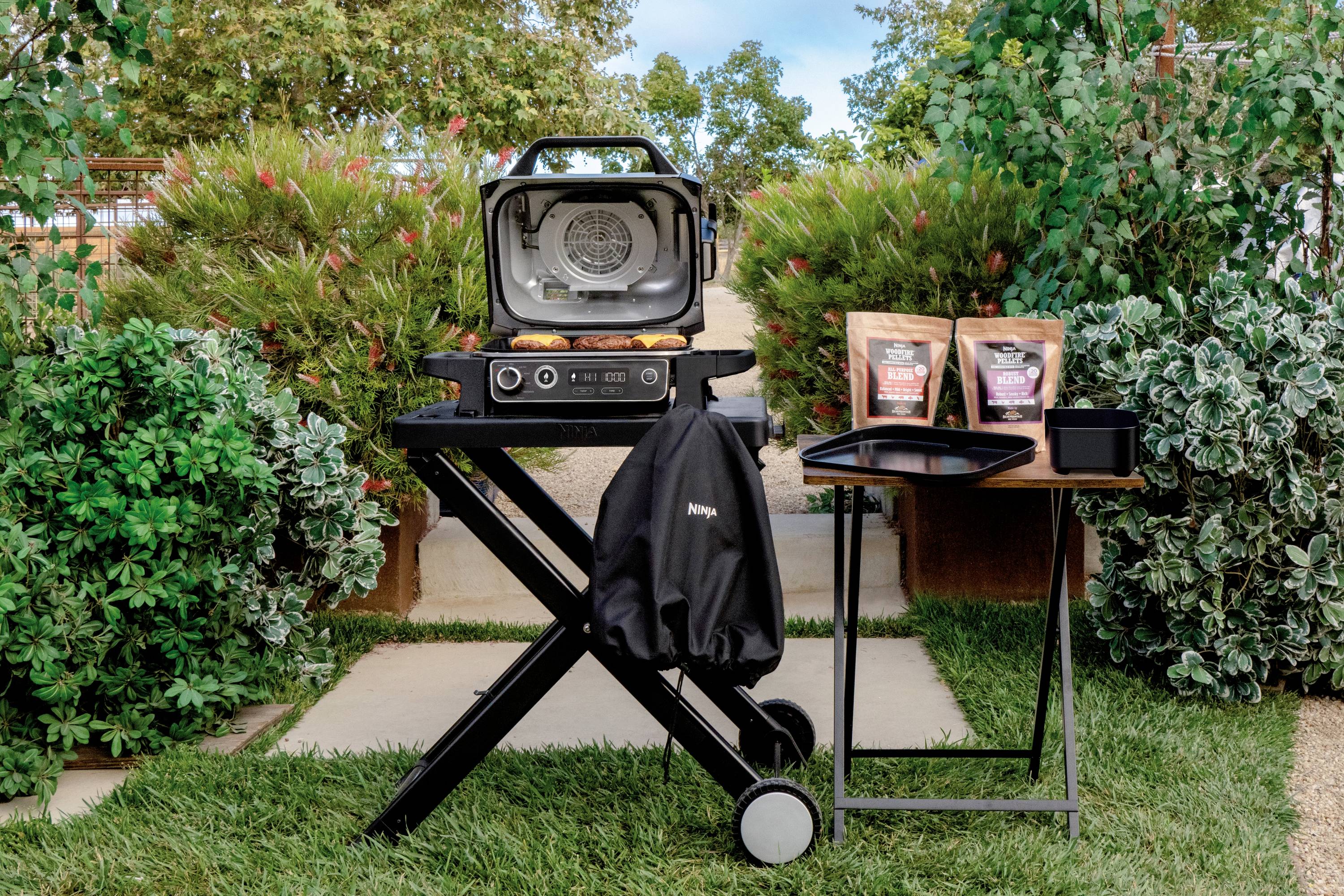A portable barbecue stands outdoors on a lawn. Nearby are barbecue accessories and a bag of charcoal. Bushes are visible in the background.