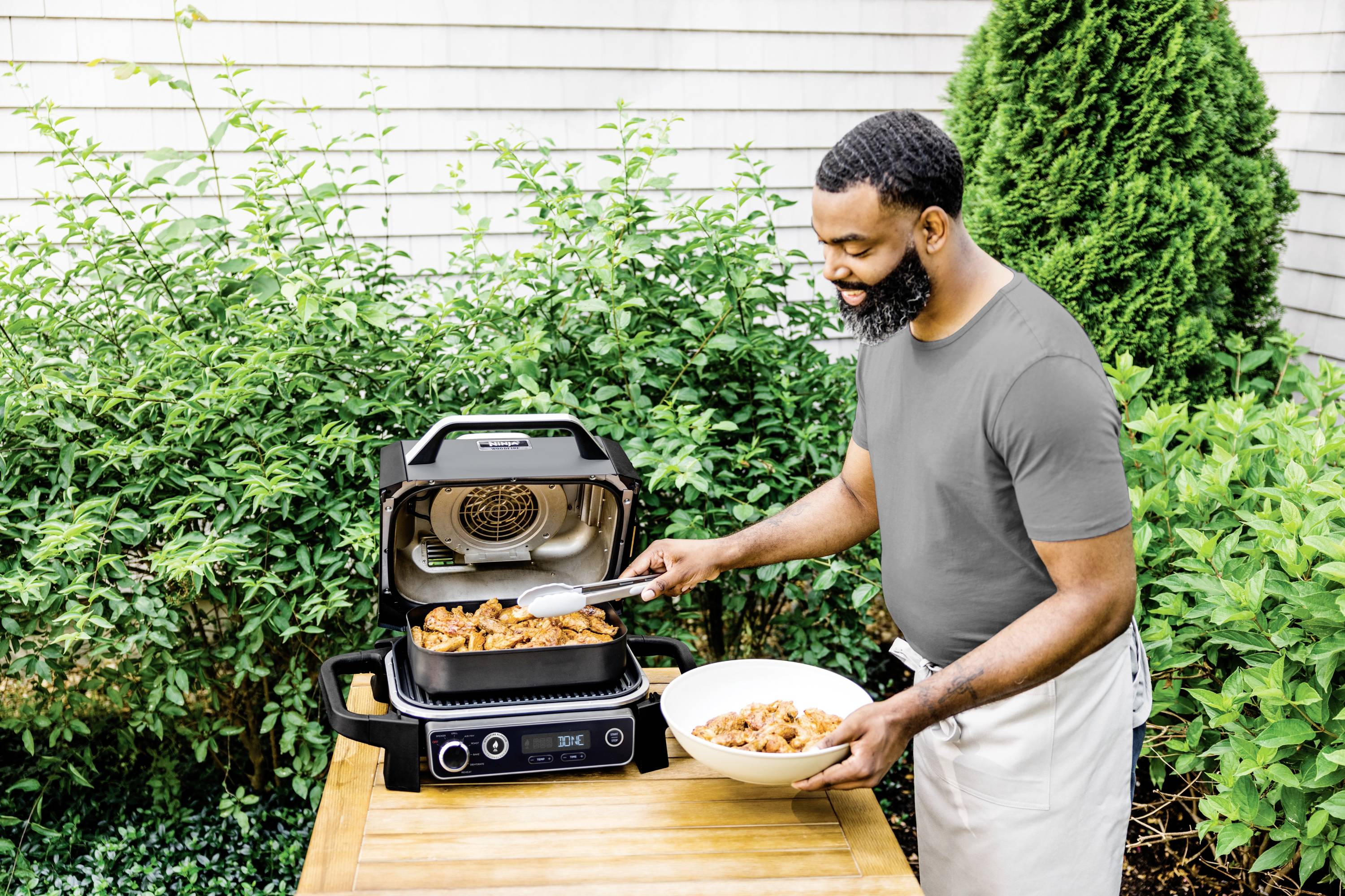A man is grilling meat on an electric barbecue in the garden and placing it in a bowl. Bushes can be seen in the background.