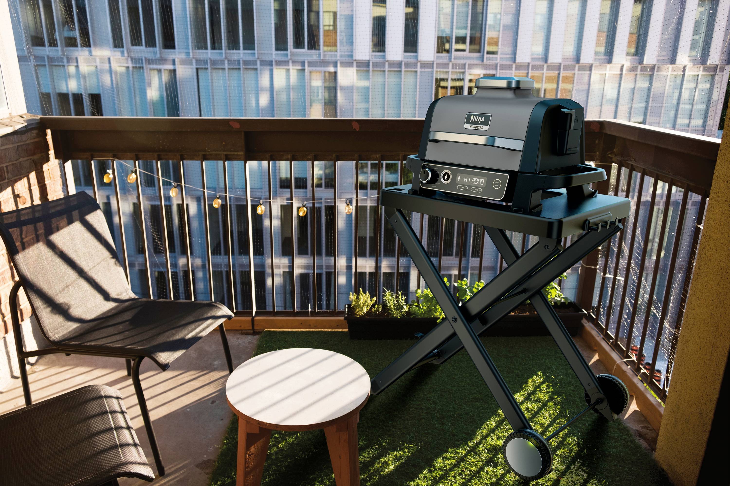 A modern barbecue stands on a small balcony overlooking an urban building. Beside it is a table and a chair.