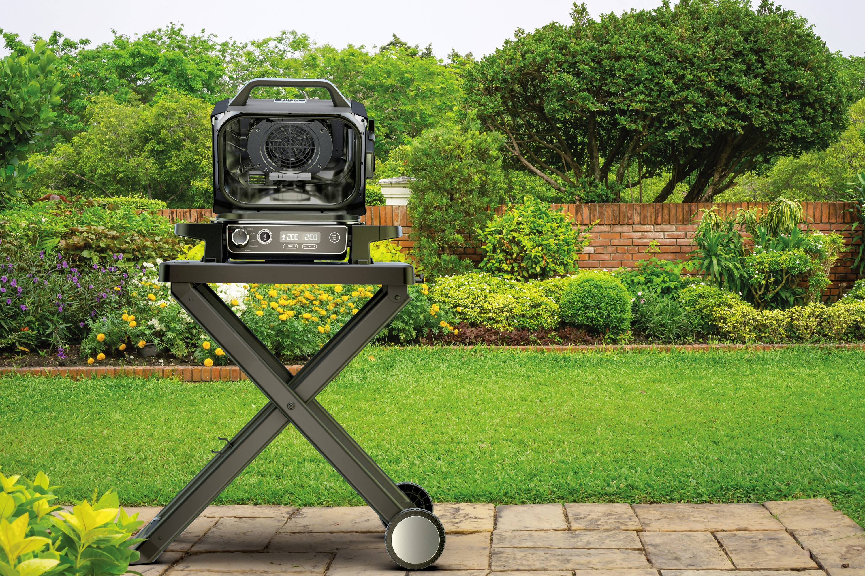A black barbecue on wheels stands on a patio. In the background, a well-maintained garden with trees and shrubs is visible.