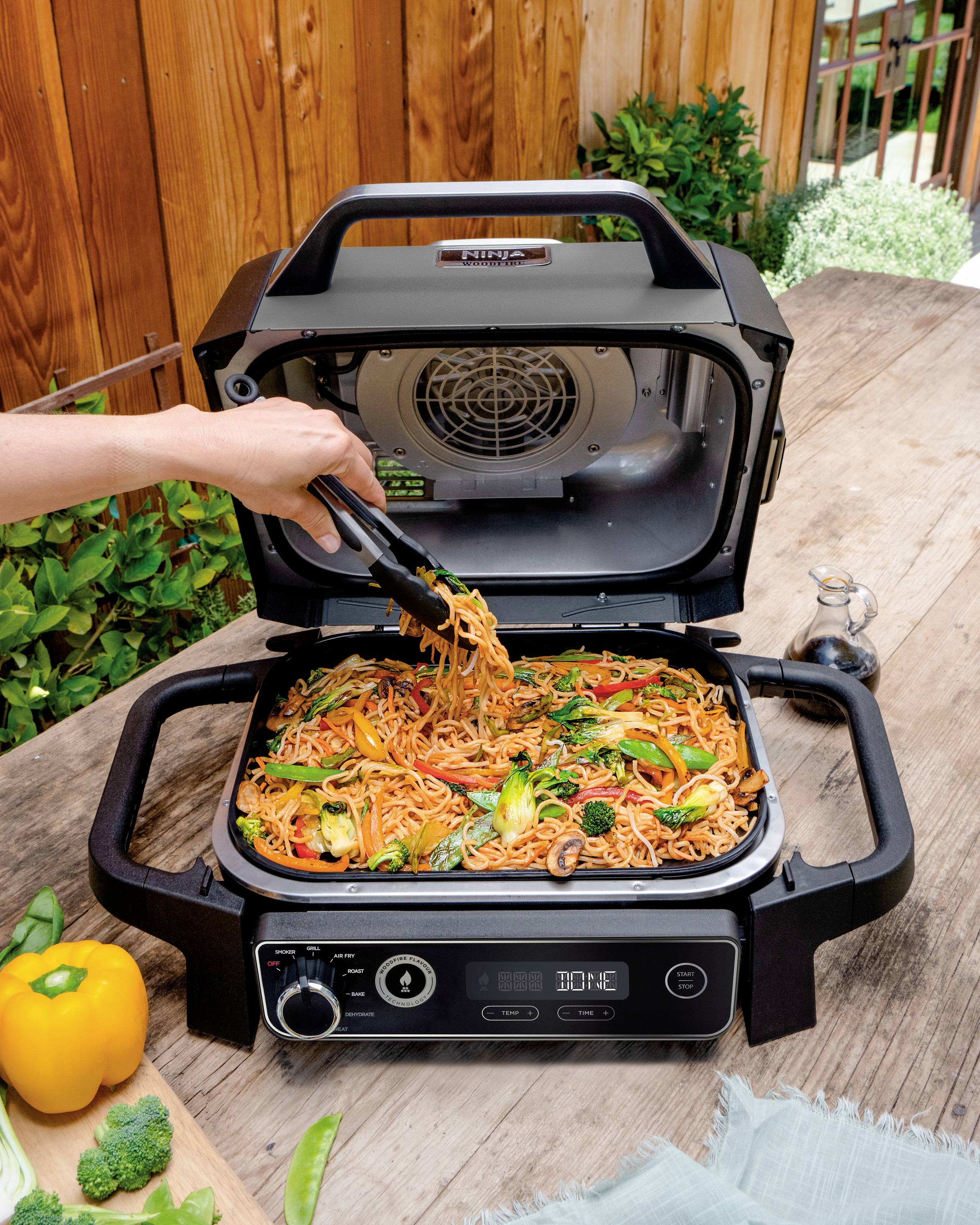 A person is lifting cooked noodles with vegetables using tongs from an open tabletop grill. A wooden fence and plants are visible in the background.
