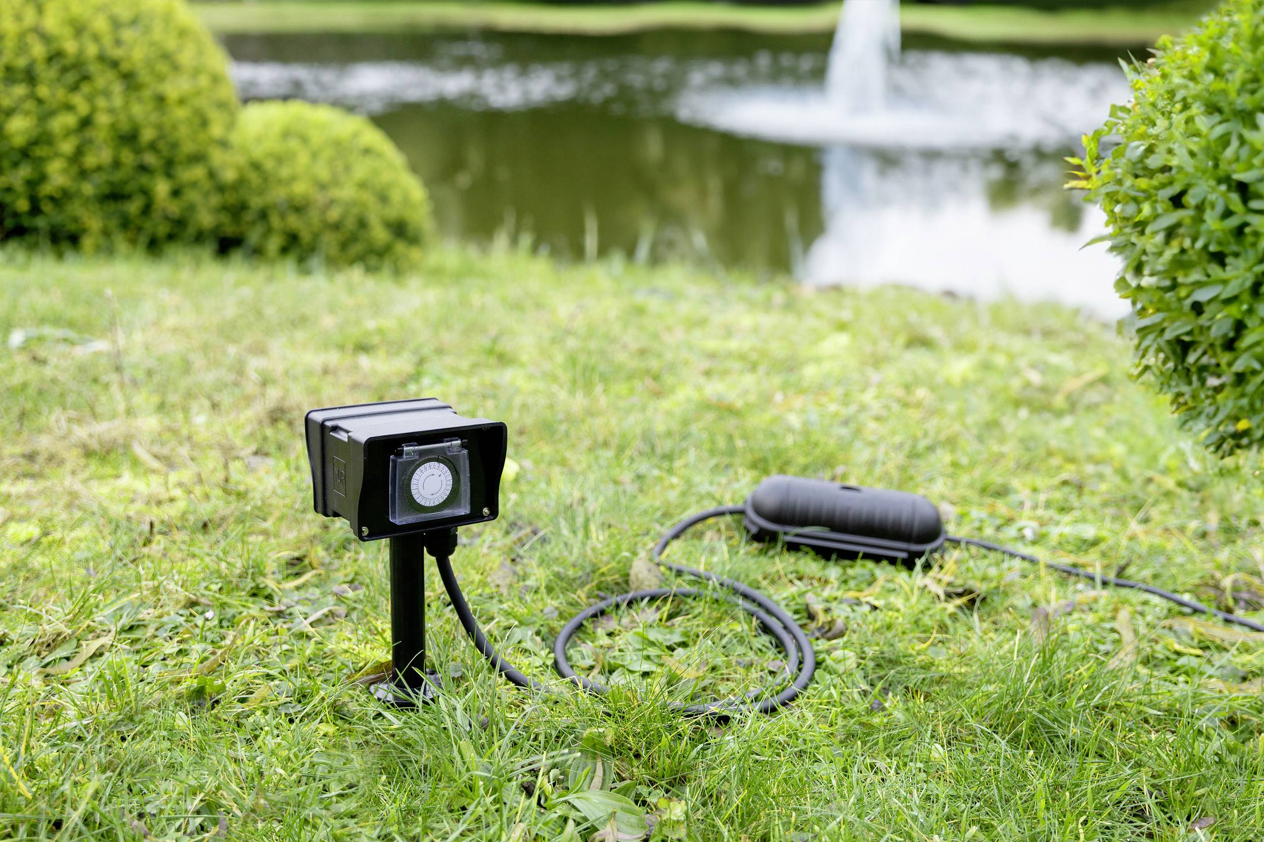 A watering sensor is installed on a lawn in front of a pond. A fountain in the background is spraying water.