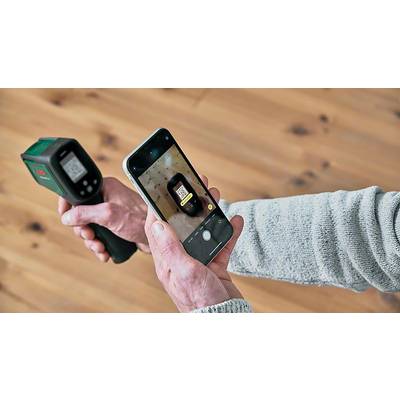 A person is holding an infrared thermometer and a smartphone, photographing the thermometer's screen. The background is a wooden floor.