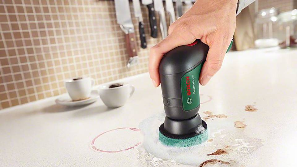 A person is using an electric power tool with a brush attachment to clean a spilled liquid on a kitchen worktop.