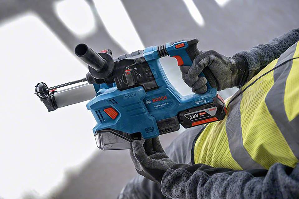 A person is holding a blue Bosch drill in their right hand, wearing a yellow high-visibility vest and work gloves.