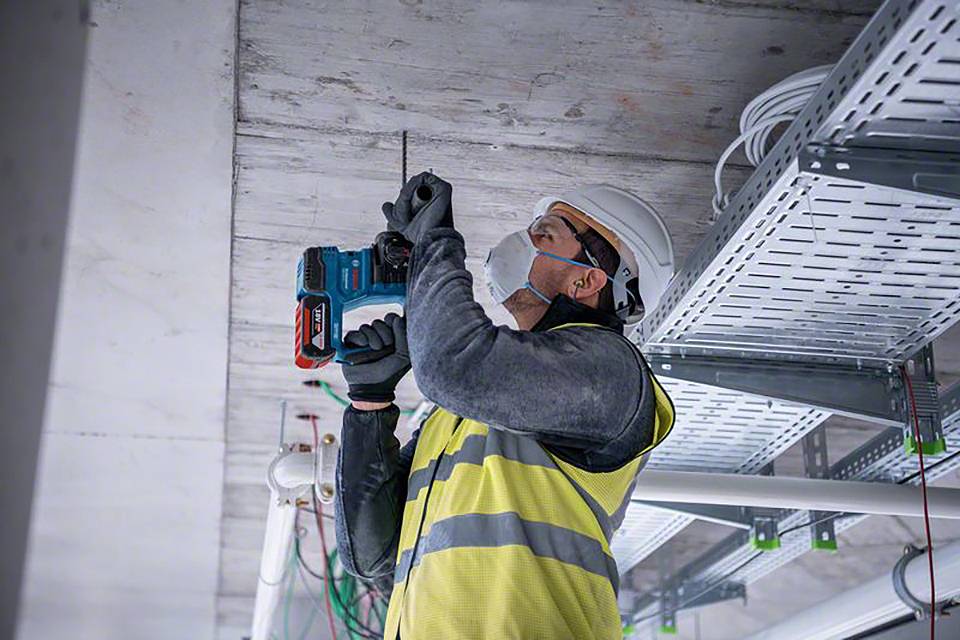 A construction worker wearing a hard hat and high-visibility vest is drilling into the ceiling of a building. Cable trays and pipes are visible in the background.