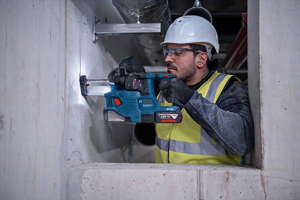 A construction worker is wearing a hard hat and safety glasses, using a drill to bore into a concrete wall, demonstrating focused work.