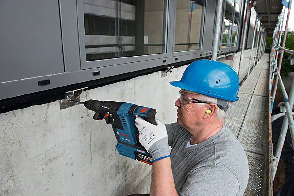 A worker in safety equipment is using a drill to secure a fitting to a concrete foundation beneath windows.
