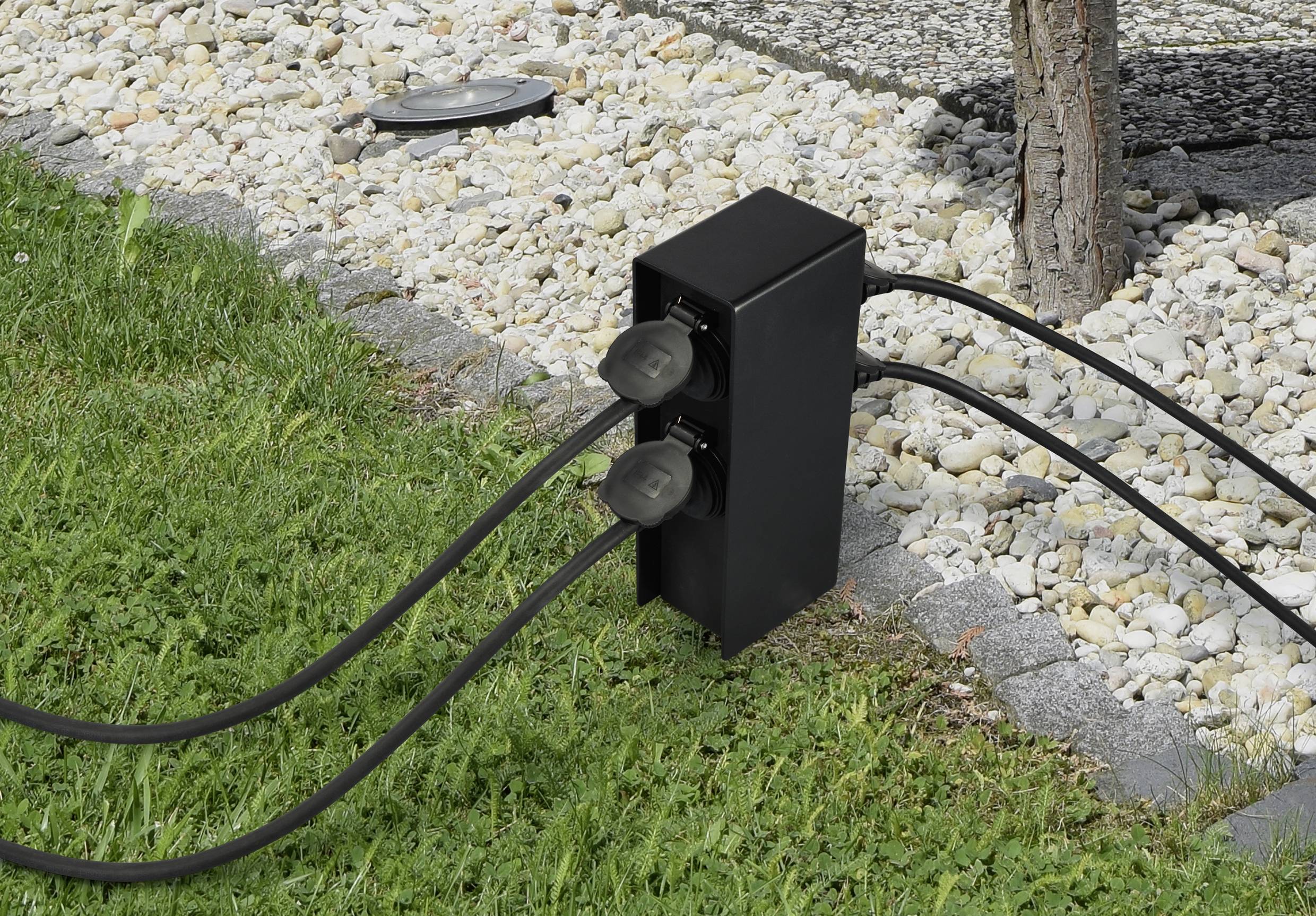 A black power strip outdoors, with three cables connected to it, is situated on a grassy area near a tree.
