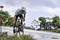 A cyclist in sports attire rides along a road in the rain, surrounded by green vegetation and a cloudy sky.