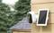 Security camera mounted on a wall with a solar panel. Trees and a roof are visible in the background.