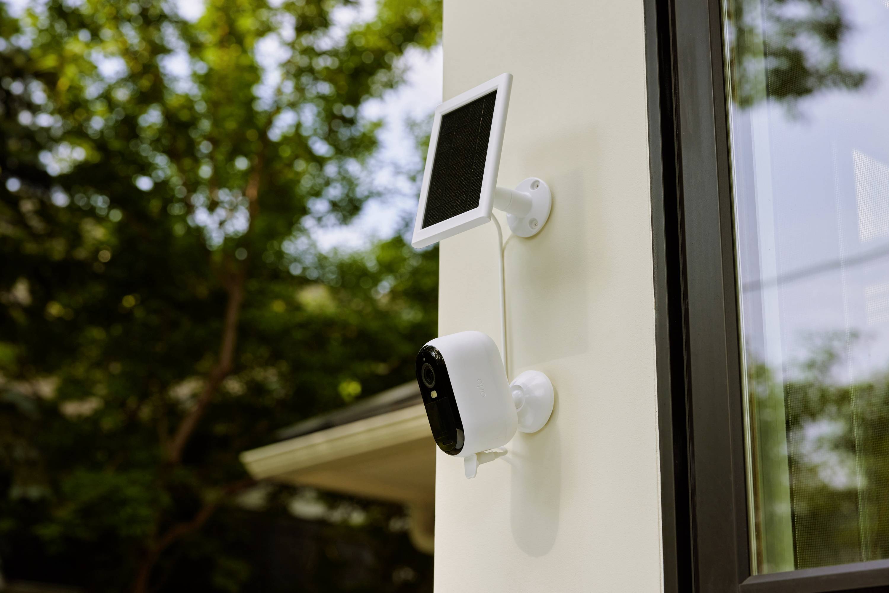 A solar-powered security camera is mounted on a house wall, surrounded by green trees in the background.