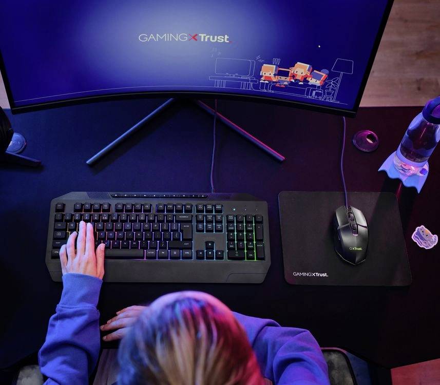 A person is playing a computer game on a monitor displaying 'Gaming Trust'. Keyboard and mouse are visible in the scene.