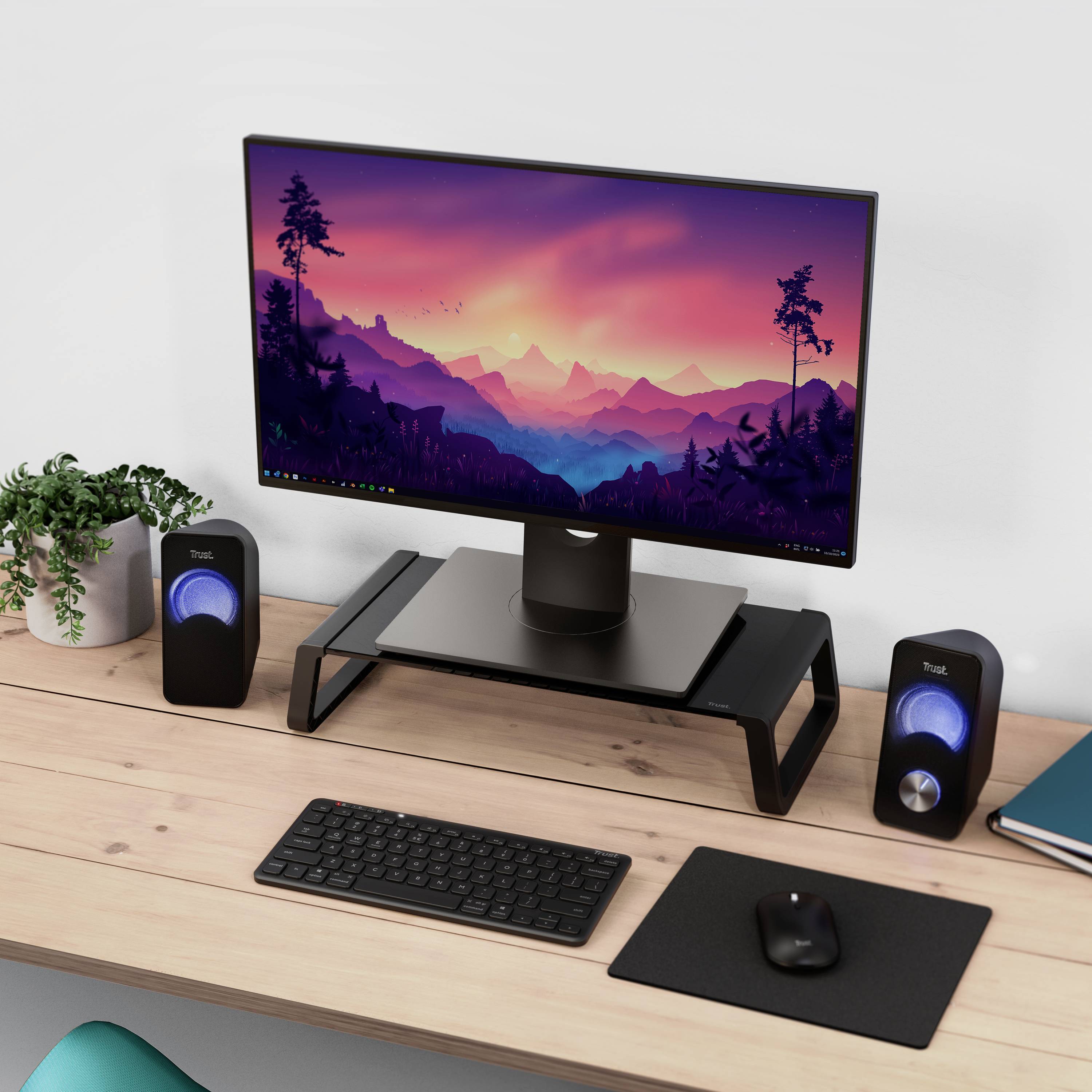 A computer screen displays a colourful mountain landscape. On a table are speakers, keyboard, mouse and plants.