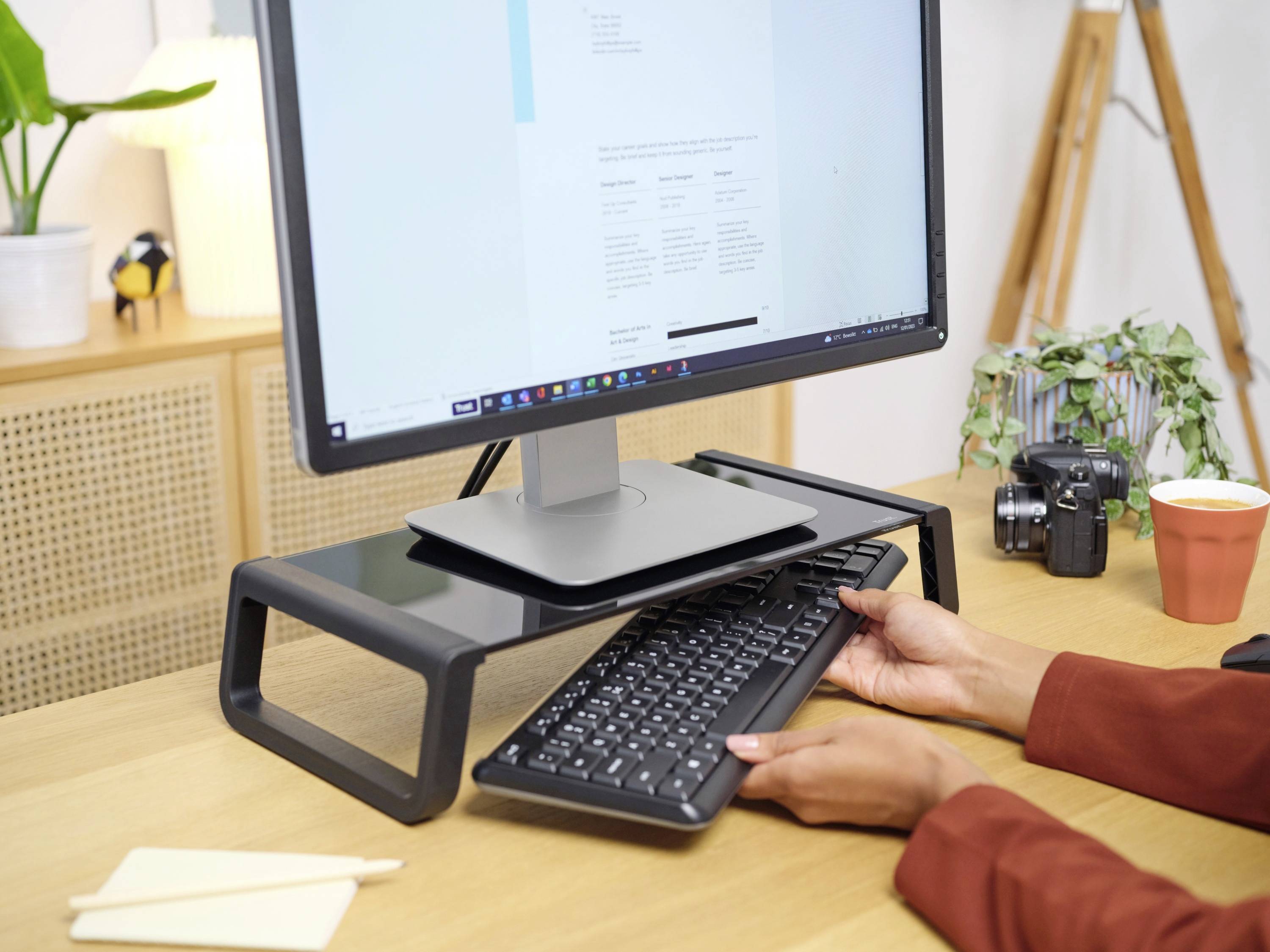 A person pulls out a keyboard from underneath a monitor on a desk. A camera and a coffee mug are sitting on the table.