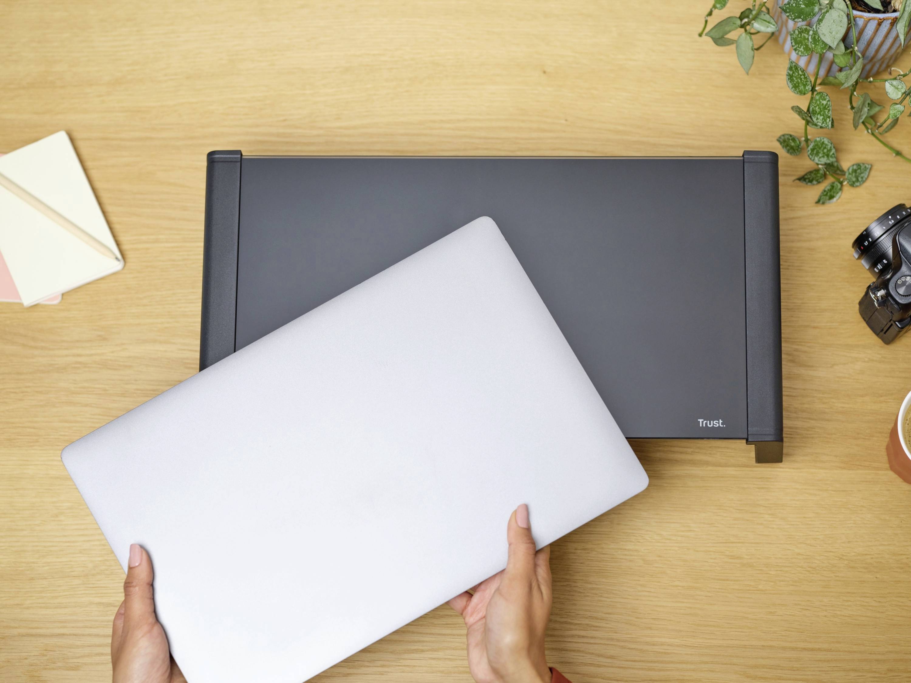 A person is holding a closed laptop over a laptop desk. A camera, plants, and notepads are lying on the table.