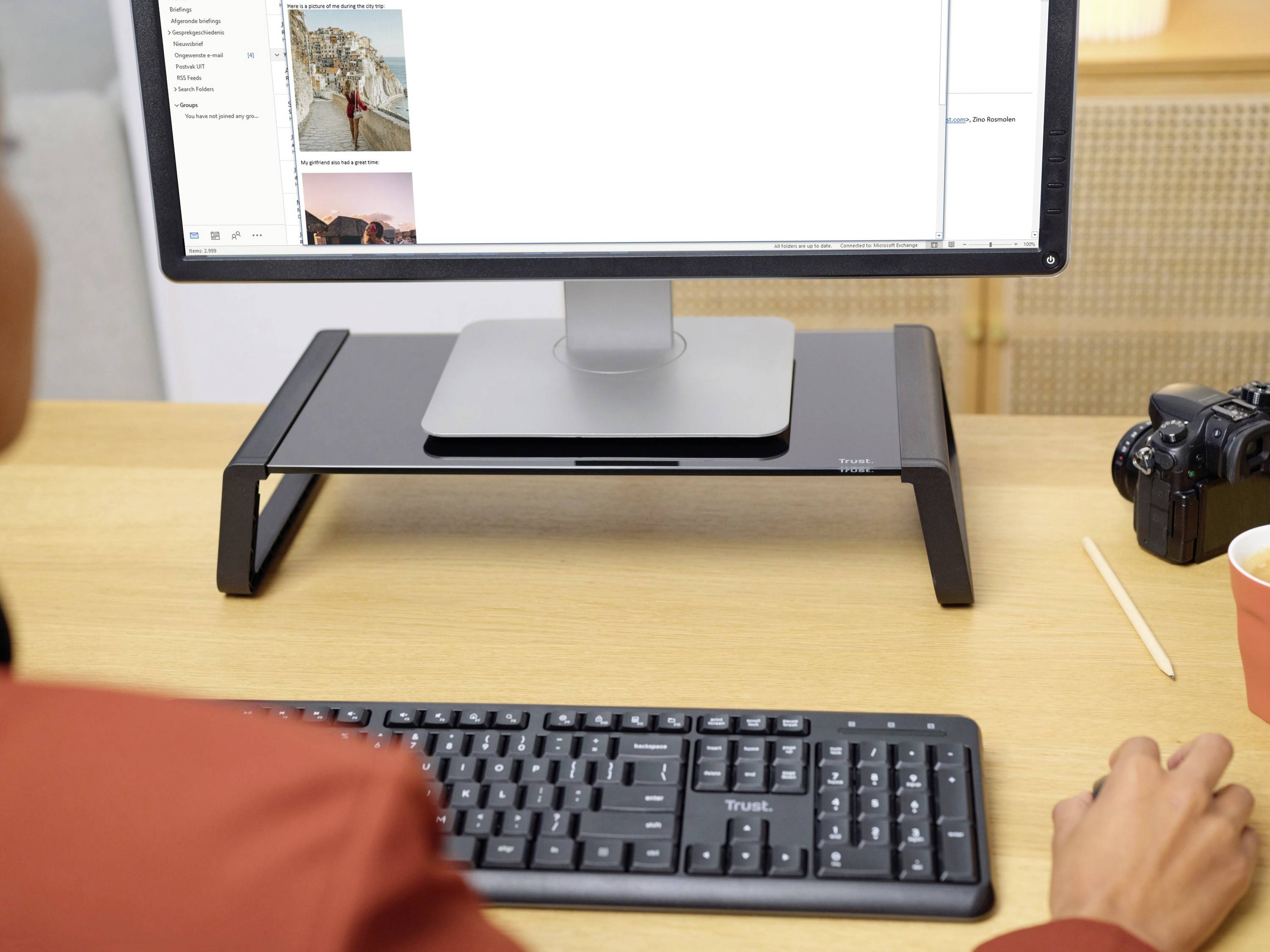 A desk with a computer monitor on a stand, a keyboard and a camera beside it. A person is working on the computer.