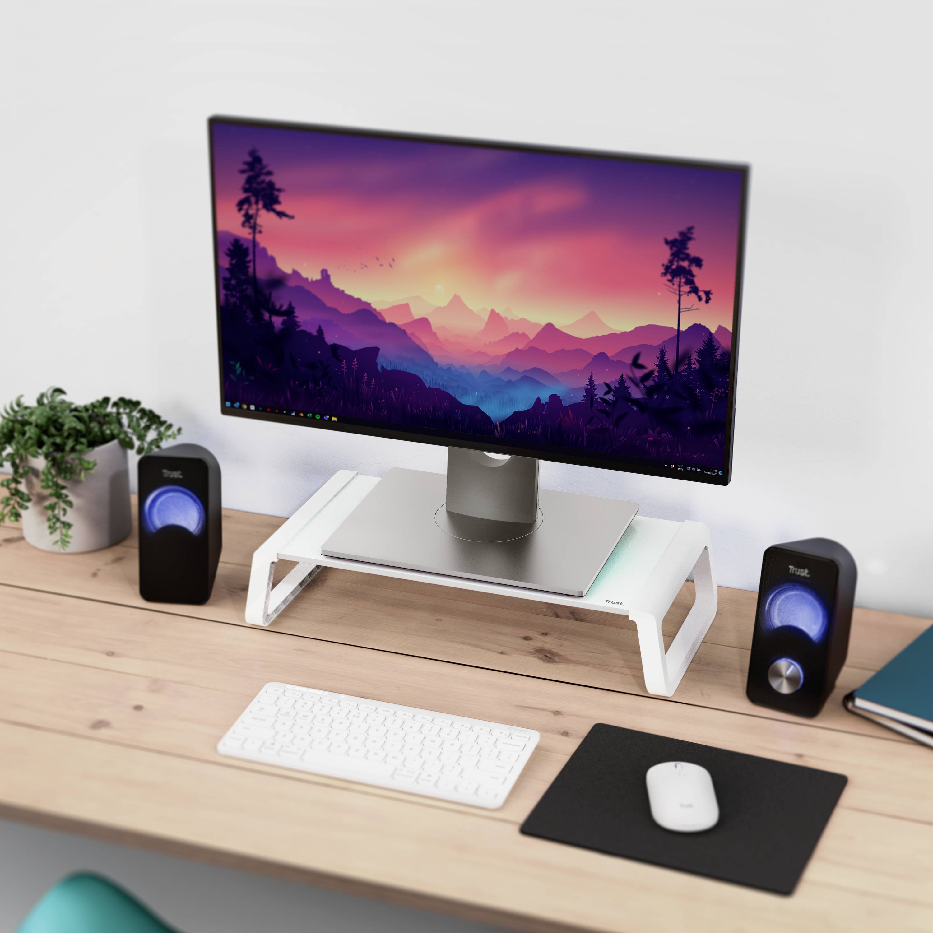 A desk with a computer screen displaying a landscape, speakers, keyboard, mouse, and a potted plant.