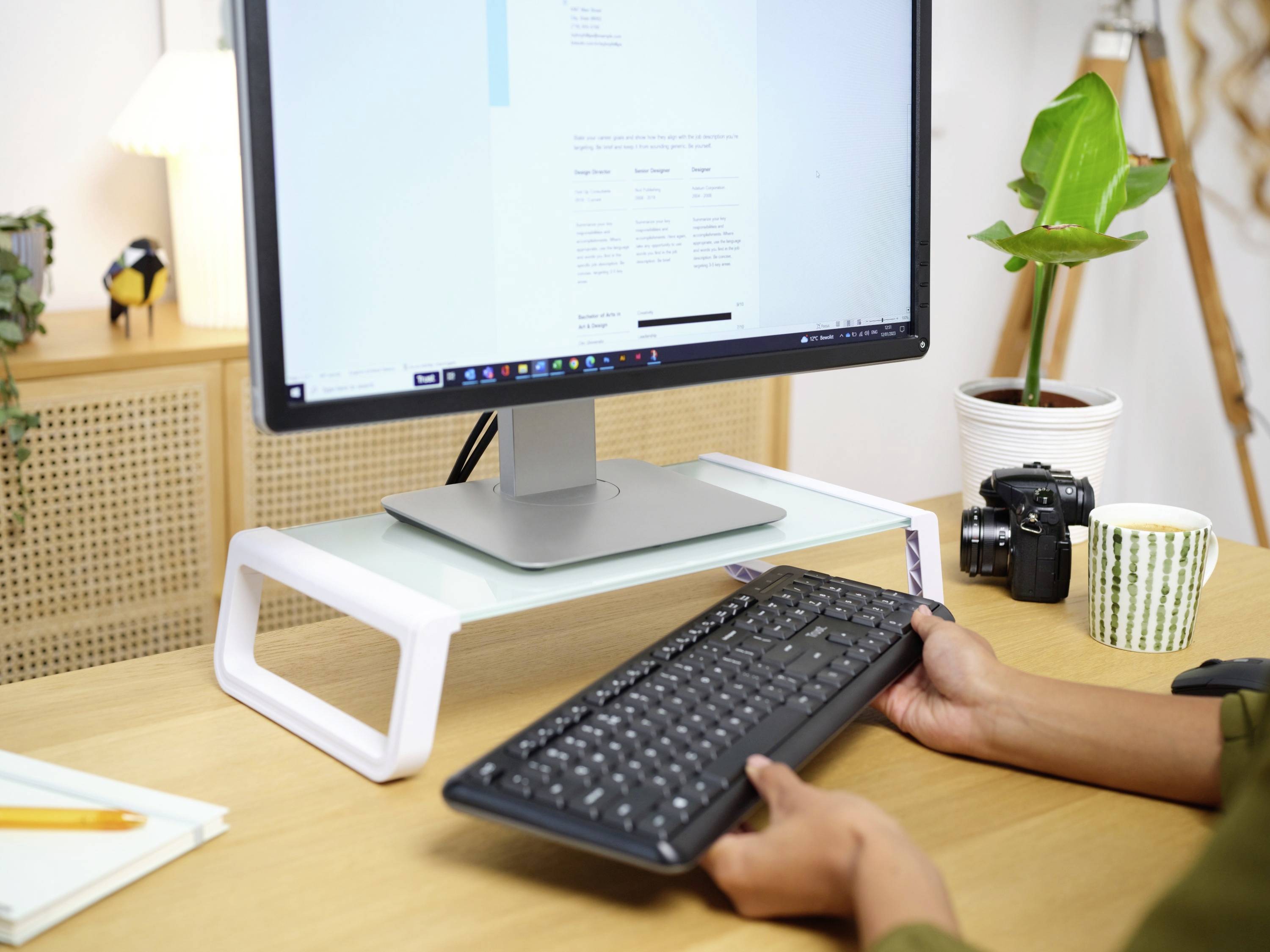 A desk with a monitor on a stand, a person holding a keyboard. A plant, camera, and mug in the background.