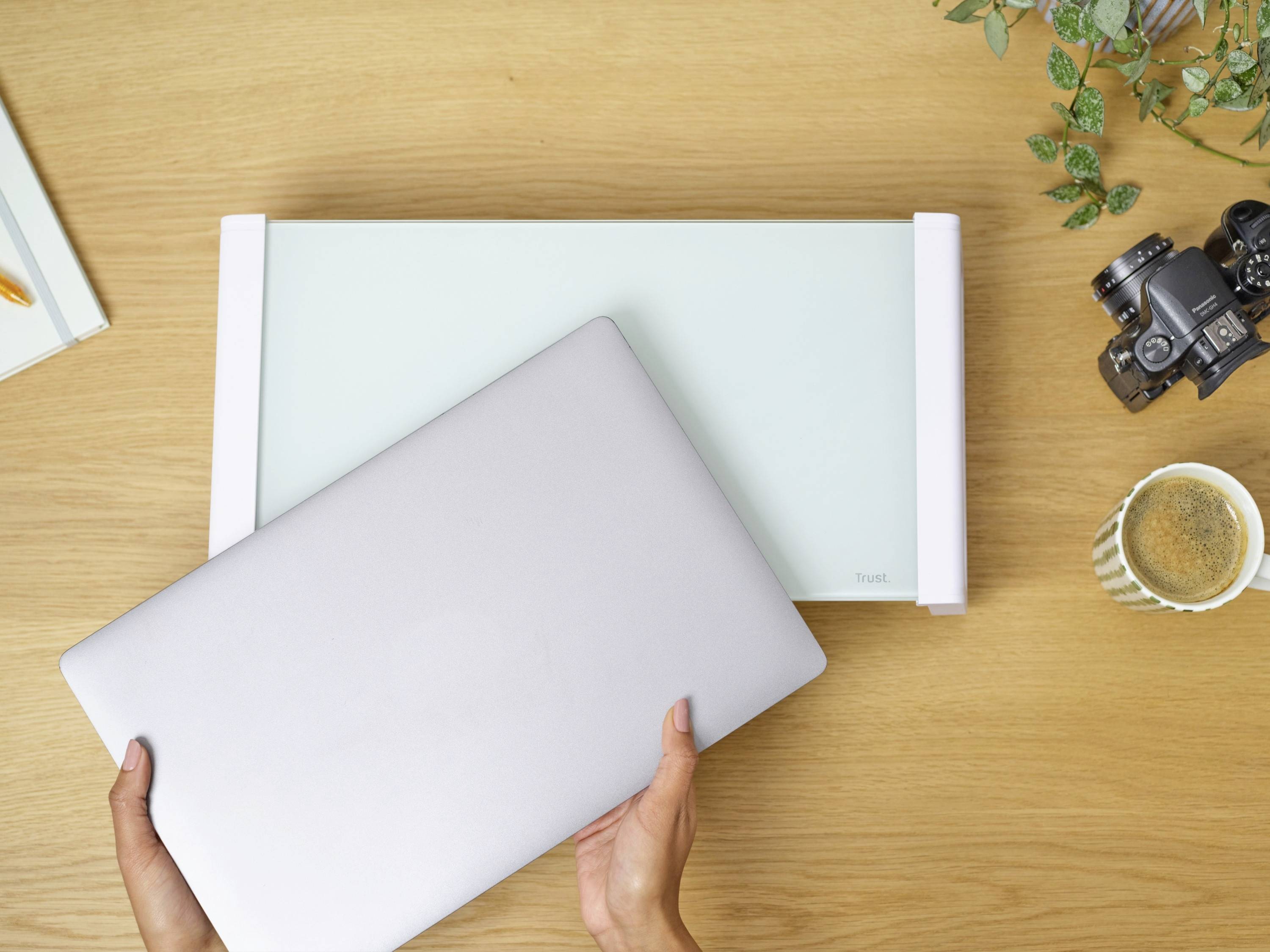 A person places a laptop on a green desk organiser on a wooden table. To the right are a camera, a plant, and a mug.