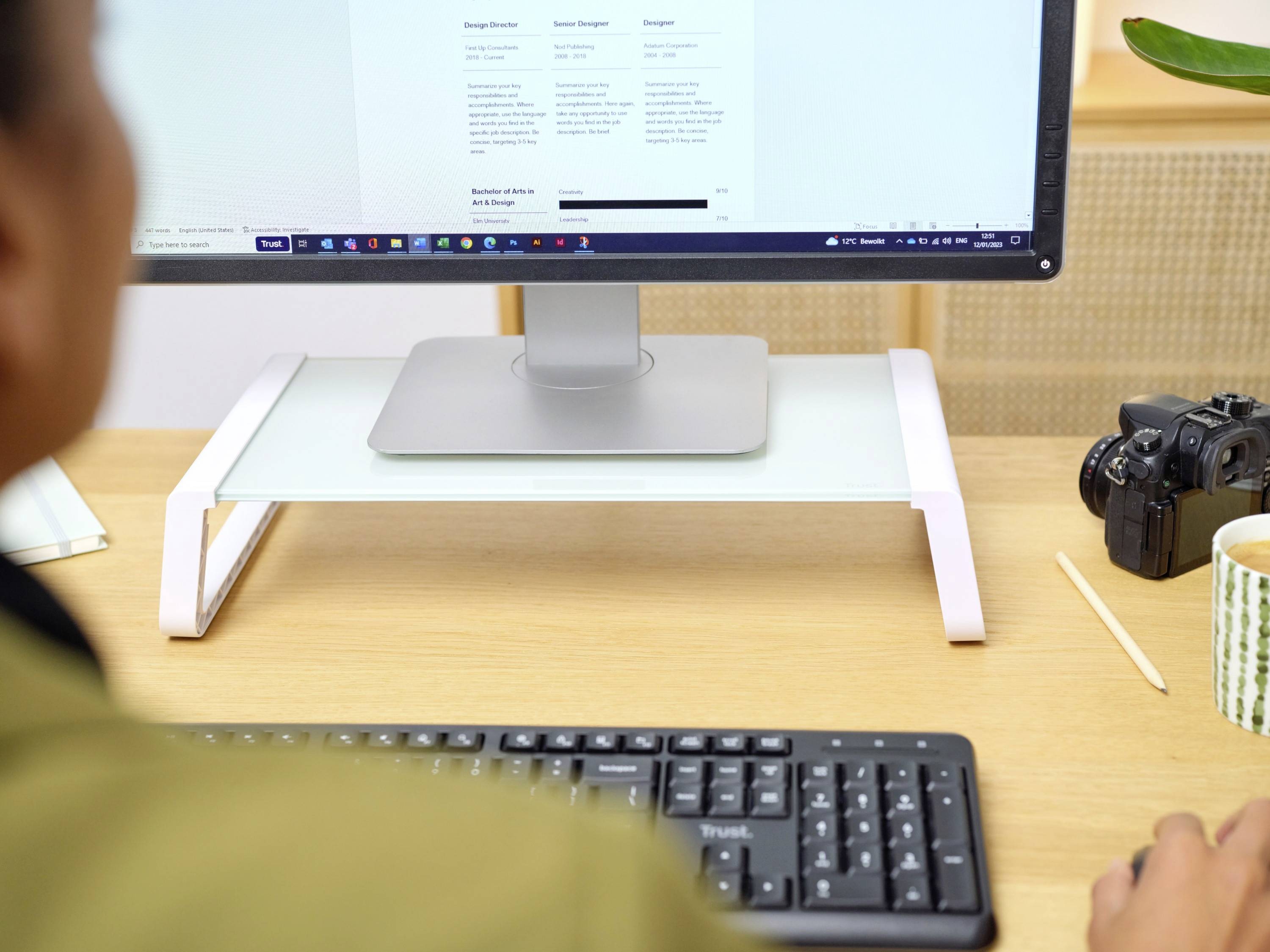 A person is sitting at a desk, looking at a monitor displaying text. On the desk are a keyboard, camera, and mug.