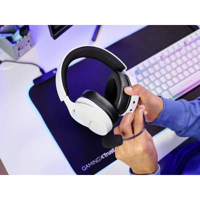 A person is holding a white gaming headset over a desk with a keyboard and mouse. A monitor is visible in the background.