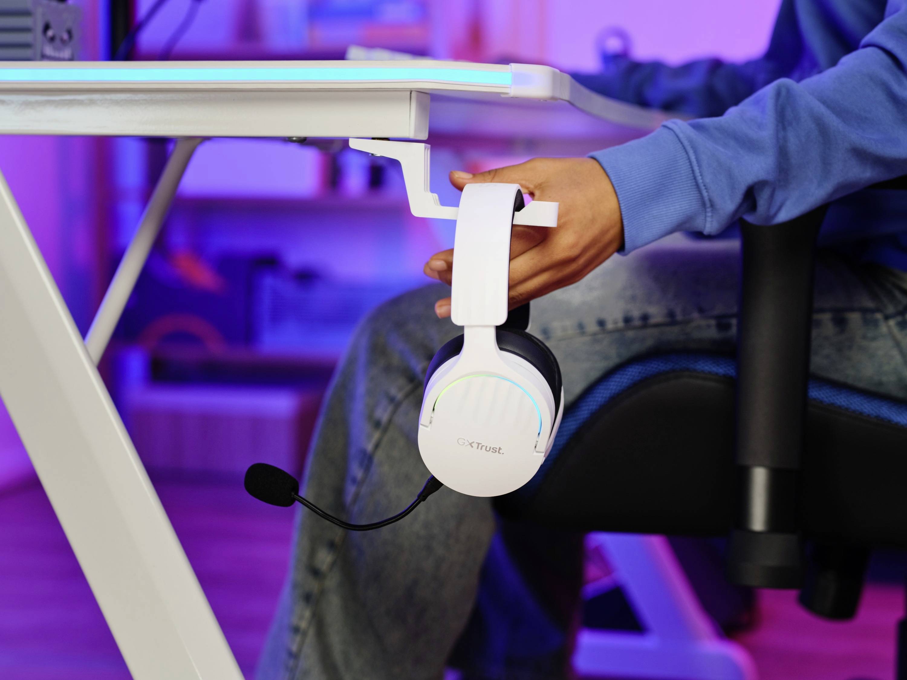 A person is hanging a white, illuminated headset on a desk mount. In the background, a desk and warm lighting are visible.