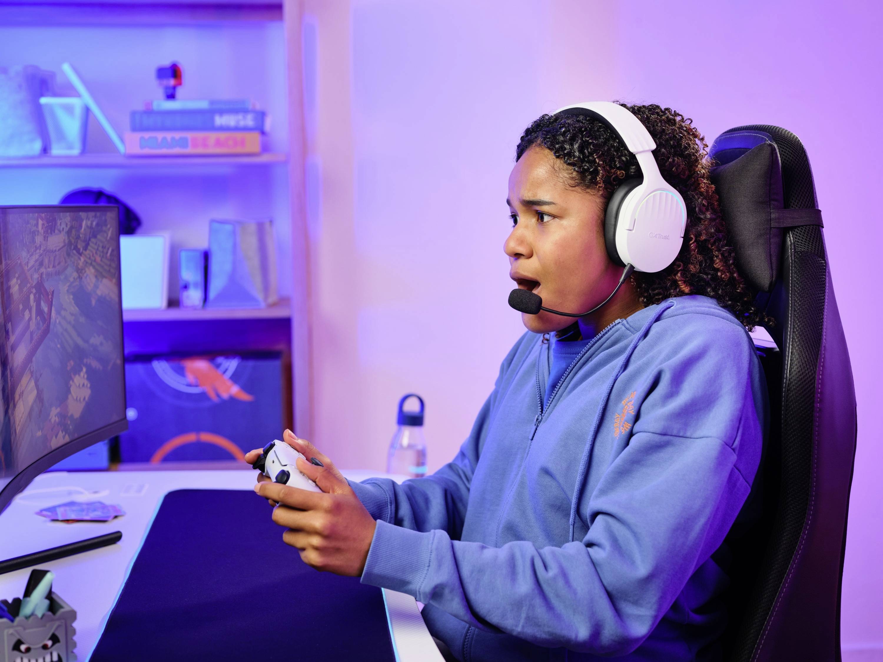 A person is sitting at a desk wearing headphones and playing a video game on a monitor. Bookshelves are visible in the background.