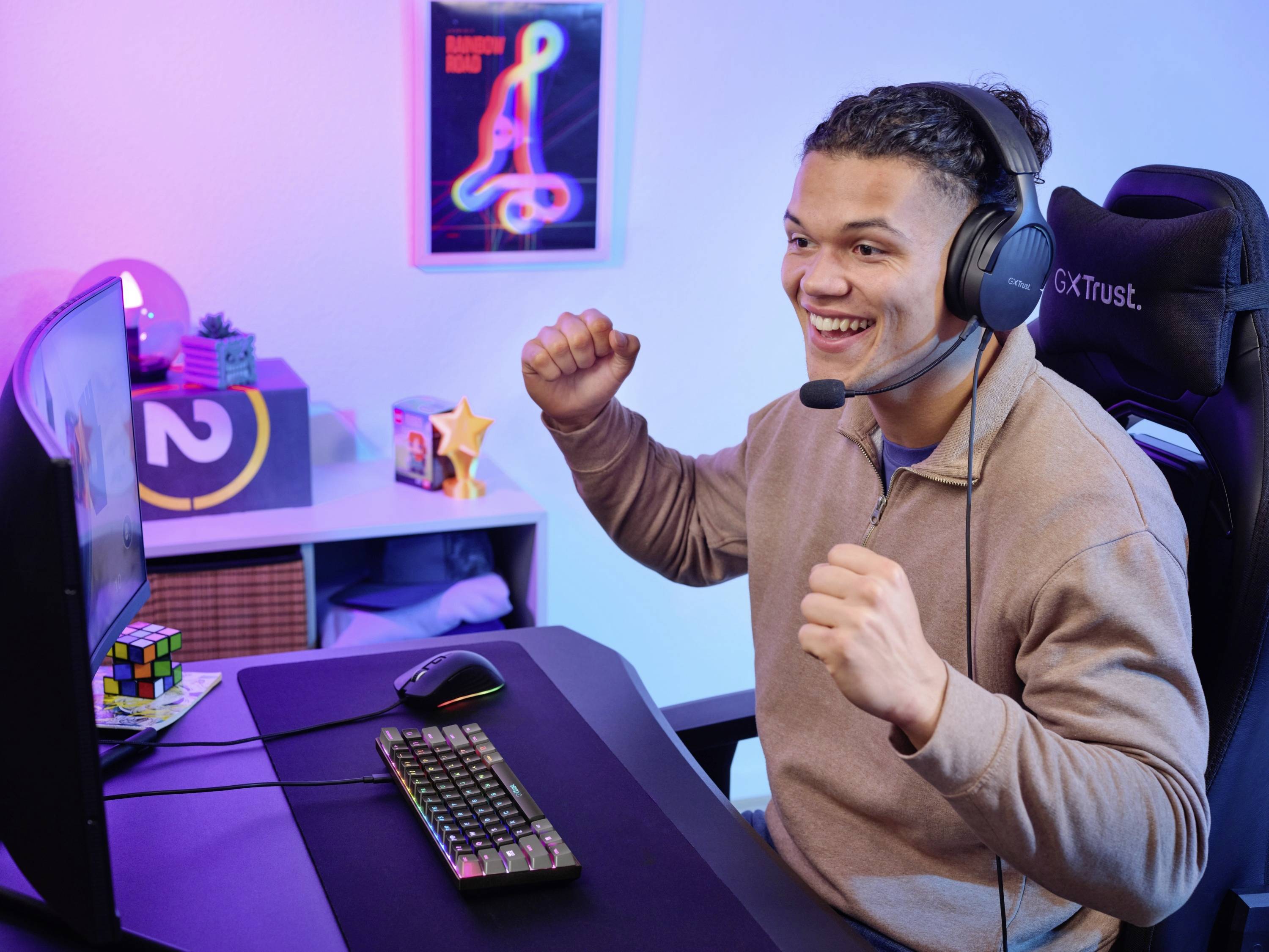 A man wearing headphones sits in front of a computer screen, looking enthusiastic. The room is illuminated with colourful lighting.