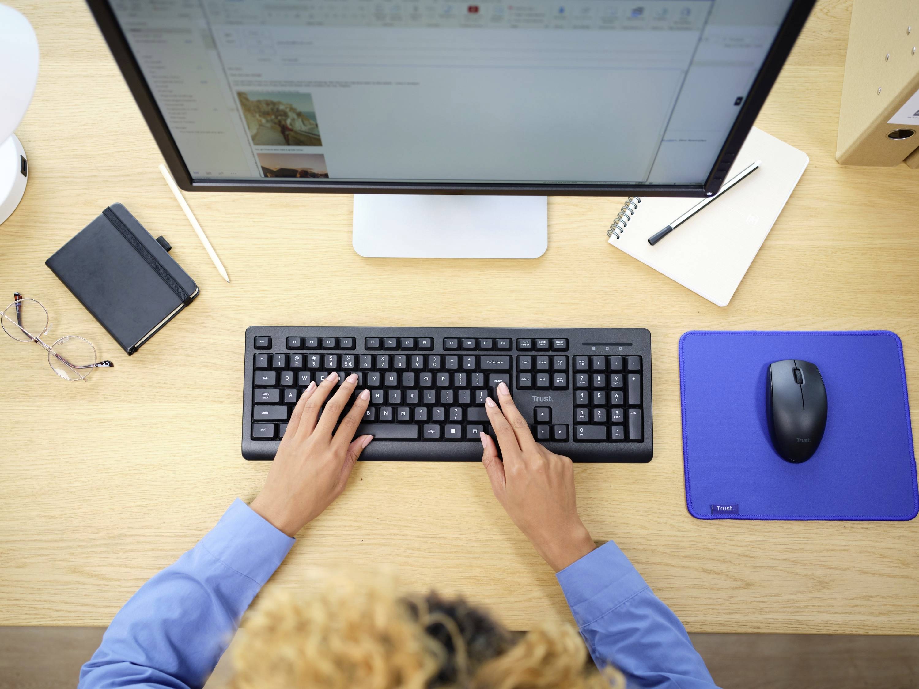 A person is typing on a black keyboard in front of a computer screen. A notebook, a pair of glasses, and a mouse are lying on the desk.