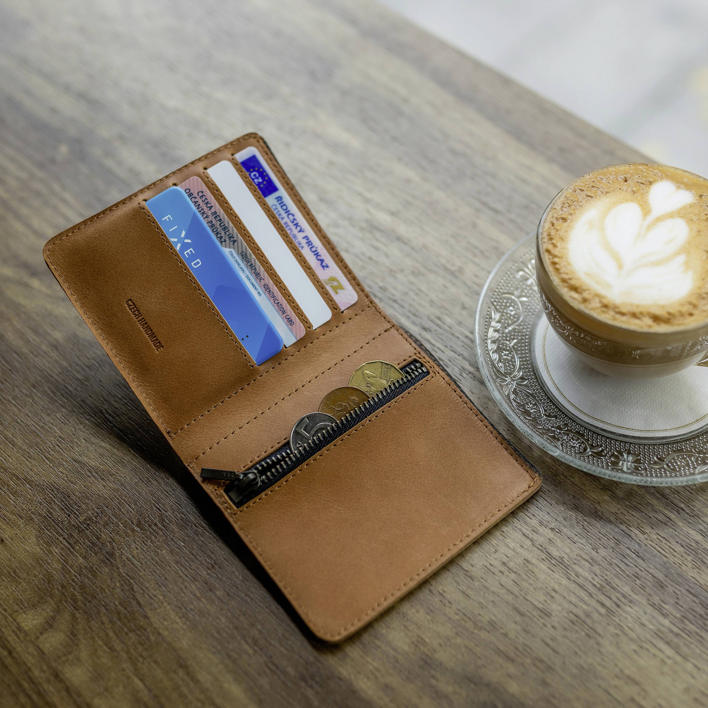 An open brown wallet with credit cards and coins beside a cup of cappuccino on a wooden table.
