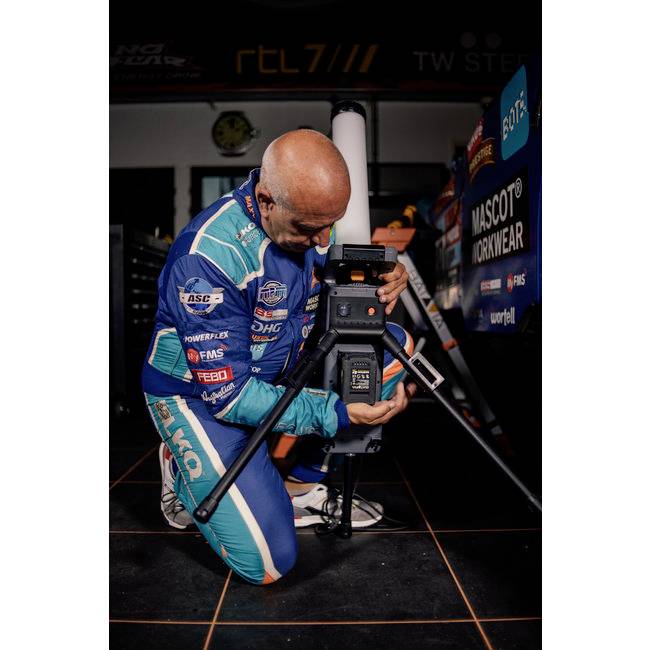 A person in a blue racing suit is kneeling beside a camera in a workshop. Sponsor logos are visible in the background.