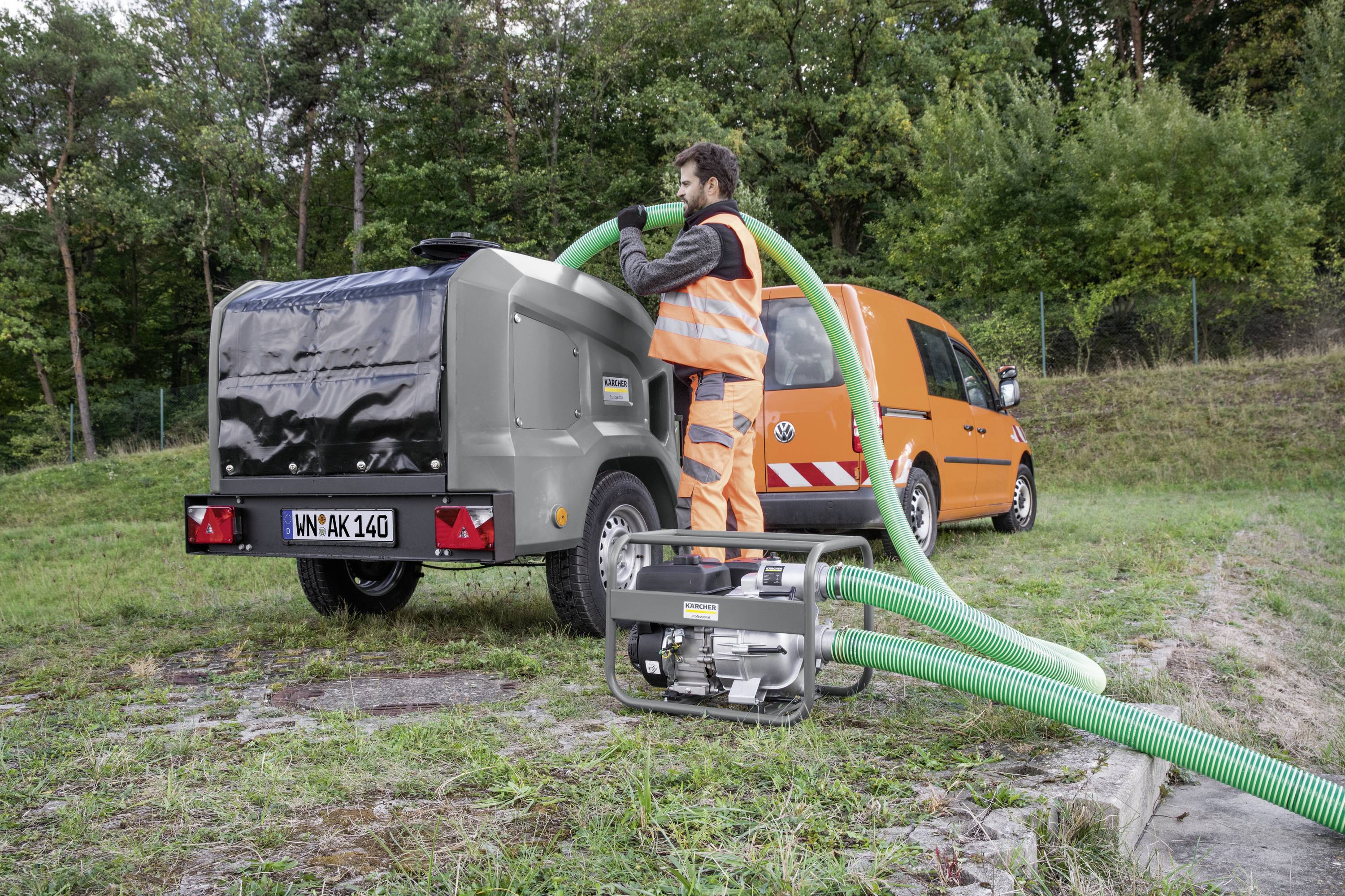 A worker in orange safety clothing is operating a hose system on a trailer next to an orange company vehicle outdoors.