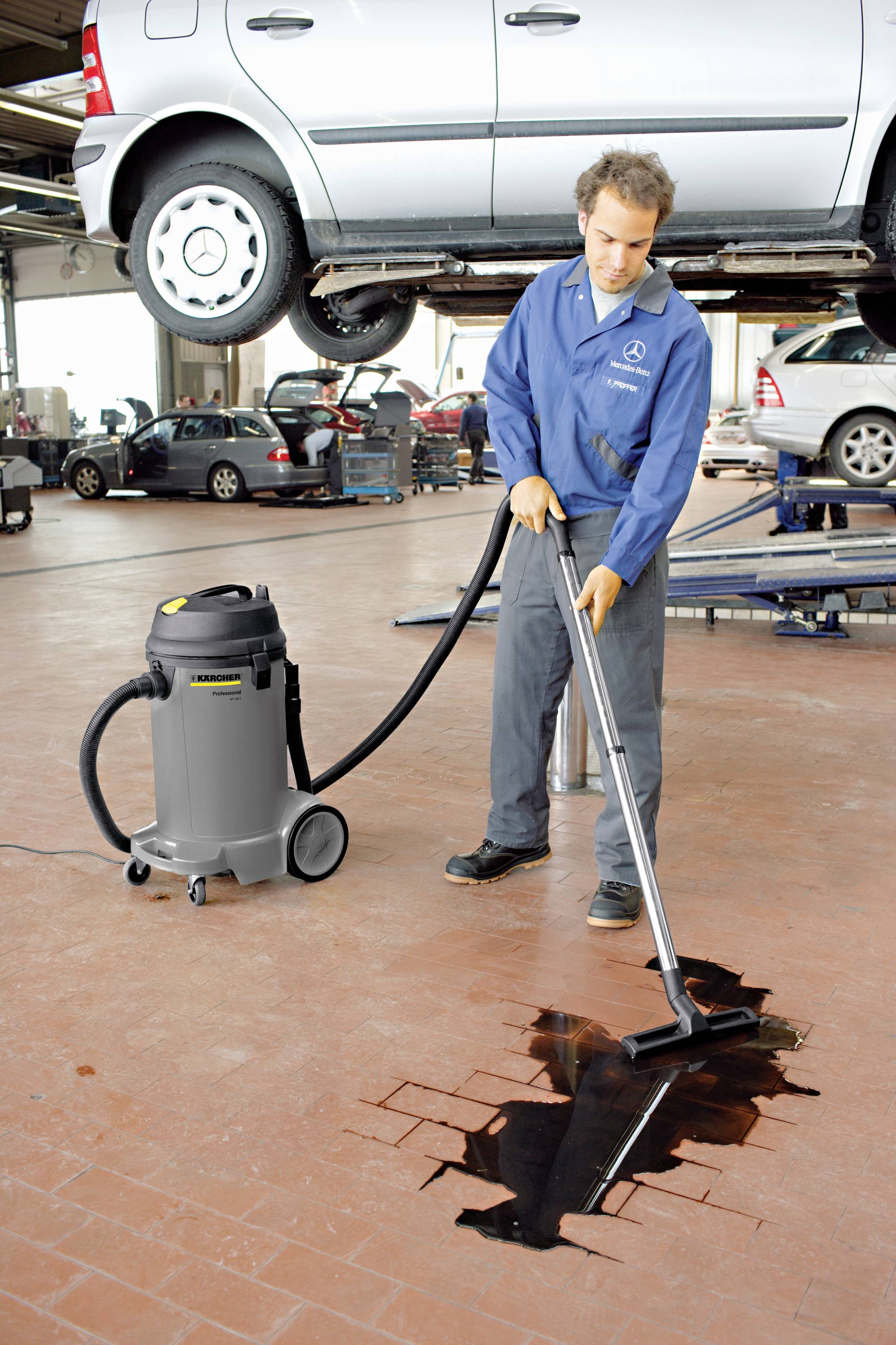 A mechanic in a car workshop is using a vacuum cleaner to suck up oil from the floor. A car is raised on a lift in the background.