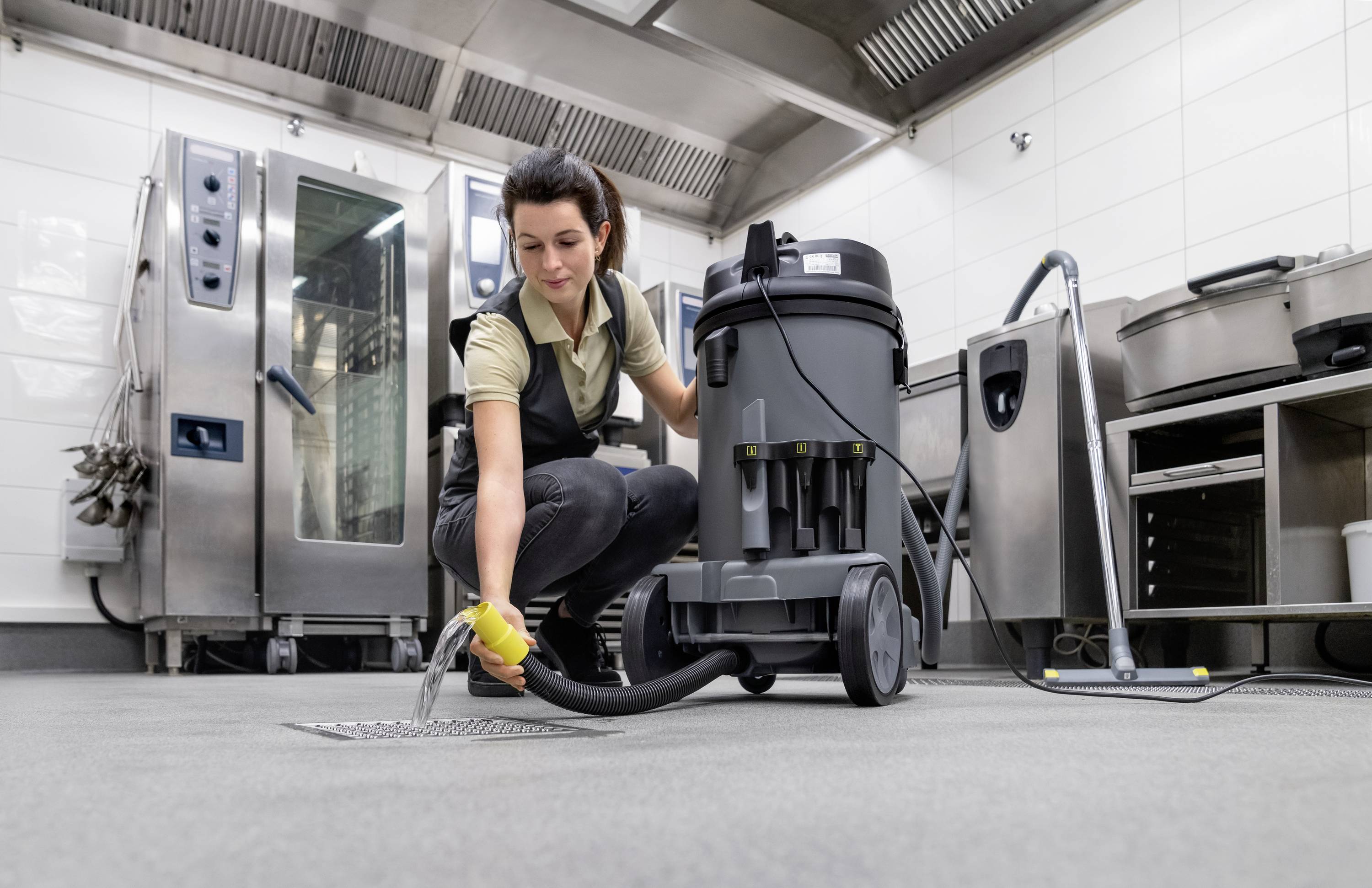A person is using an industrial vacuum cleaner to clean the floor in a professional kitchen. Kitchen equipment is visible in the background.
