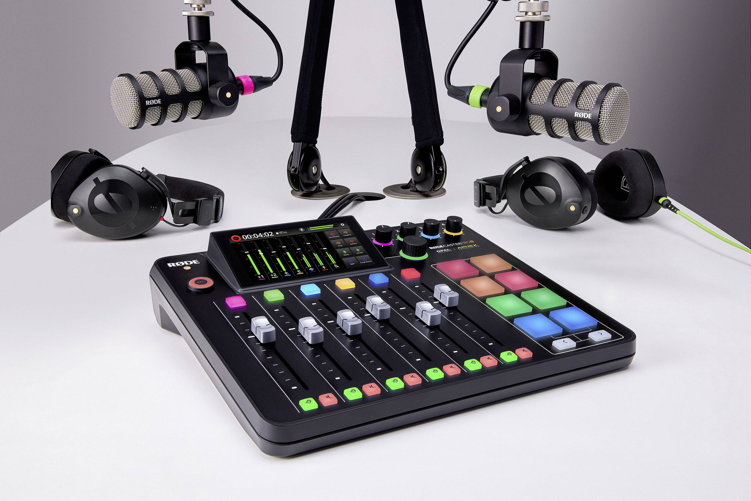An audio mixing desk with faders and colourful pads sits on a table, flanked by two microphones, headphones, and cables.